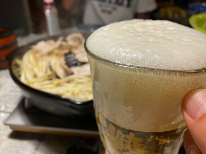 泡がきめ細かいビールのグラスと奥に見える鍋